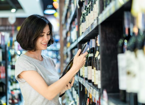 Asian beautiful woman choosing nice red wine bottle picking up from alcohol shelf in supermarket